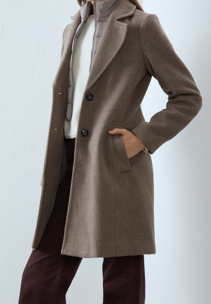Femme portant un manteau en laine taupe sur une veste grise matelassée et un haut blanc, une main dans la poche du manteau, sur un fond uni.