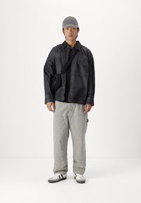 Camicia in denim nera, t-shirt nera sotto, pantaloni cargo grigio chiaro, cappellino grigio e sneakers bianche con strisce nere. Outfit casual.