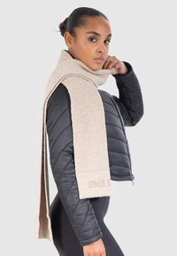 Polstret sort jakke med høj krave, parret med et beige ribbestrikket tørklæde med et logo. Tørklædet hænger over skulderen.