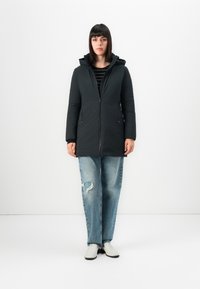 Frau in schwarzer Kapuzenjacke, gestreiftem Oberteil, zerrissenen hellblauen Jeans und weißen Schuhen, die vor einem schlichten weißen Hintergrund steht.