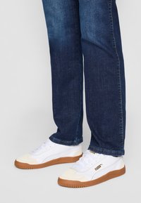 Baskets blanches avec semelles en caoutchouc couleur gomme, munies d'un laçage, d'un col rembourré et d'accents de logo dorés. Portées avec un jean en denim bleu foncé.