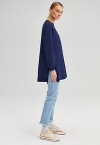 Top a maniche lunghe blu navy con una vestibilità ampia, caratterizzato da dettagli arricciati sui lati. Abbinato a jeans azzurri strappati e sneakers beige.