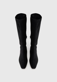 Botas pretas acima do joelho em camurça macia. Bico quadrado e cano alto com costuras visíveis e zíperes laterais para facilitar o uso.