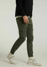 Hombre vistiendo pantalones cargo verde oliva, zapatillas blancas, calcetines beige, chaqueta negra y camisa a rayas, caminando, visto desde la mitad del muslo hacia abajo.