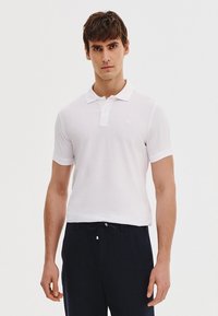 Biała koszulka polo z klasycznym kołnierzykiem i krótkim rękawem, wykonana z gładkiego materiału, noszona z granatowymi spodniami. Dyskretny logo na piersi.