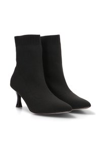 Botas de calcetín negras de punto con puntas afiladas y tacones delgados de altura media sobre un fondo blanco.