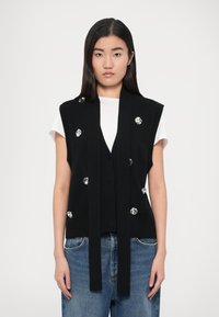 Chaleco negro de punto con abertura frontal, adornado con grandes detalles plateados. Combinado con una camiseta blanca y unos vaqueros de mezclilla.