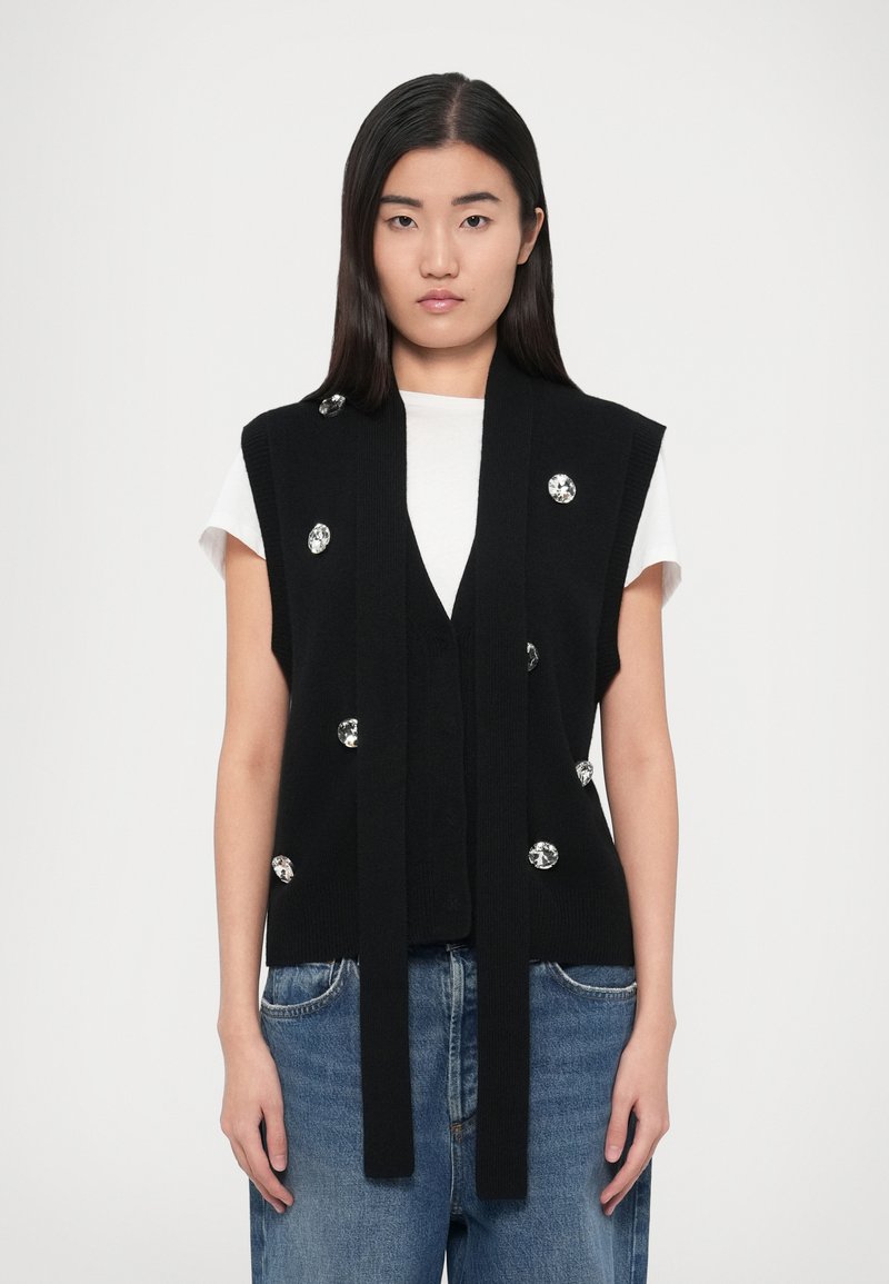 Chaleco negro de punto con abertura frontal, adornado con grandes detalles plateados. Combinado con una camiseta blanca y unos vaqueros de mezclilla.