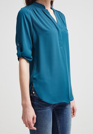 Mujer con blusa de manga larga color verde azulado con las mangas remangadas y jeans azul oscuro, de pie frente a un fondo claro y liso.