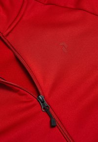 Close-up van een rood jack met zwarte rits en subtiel geperst logo op een gladde stofoppervlakte.