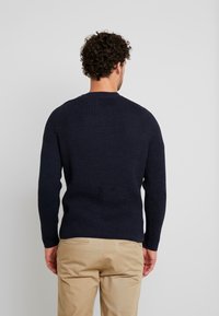 Maglione a coste navy con maniche lunghe e scollatura a girocollo, caratterizzato da un design a maglia testurizzata. Indossato con pantaloni beige chiaro.