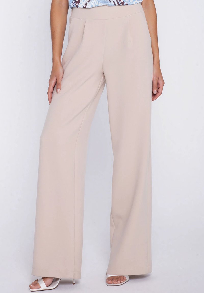 Beżowe spodnie o szerokich nogawkach noszone z białymi sandałami na obcasie z odkrytymi palcami, zestawione z wzorzystą, włożoną do środka bluzką.