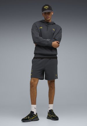 Giovane uomo in piedi con le braccia incrociate, indossa una felpa nera con cappuccio Puma, pantaloncini, cappellino, calzini bianchi e sneakers Puma nere e gialle, su sfondo grigio.