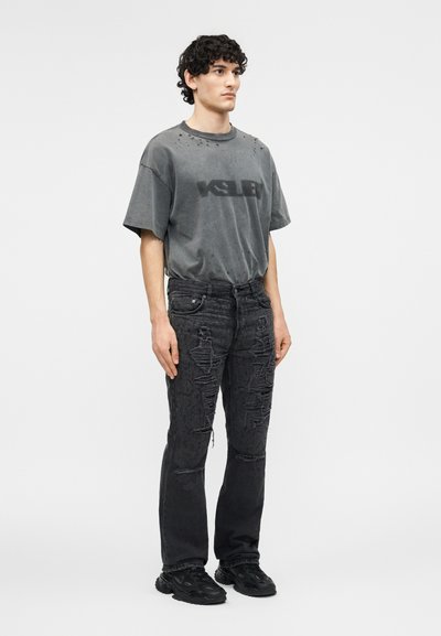 Νεαρός άνδρας στέκεται φορώντας φθαρμένη σκουρόγρια μπλούζα και πολύ ταλαιπωρημένο μαύρο τζιν με μαύρα αθλητικά παπούτσια σε λευκό φόντο.