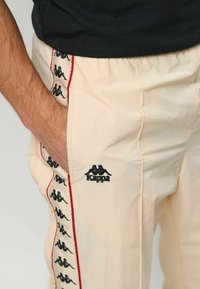 Kappa bézs nadrág fekete logóval és ismétlődő fekete mintákkal az oldalán, piros kontrasztos csíkkal és texturált anyaggal.