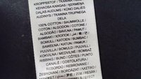 Etykieta wykonana z jasnej tkaniny, zawierająca tekst w wielu językach. Wymienia 100% bawełny z różnymi terminami dotyczącymi tkaniny wyróżnionymi pogrubieniem.