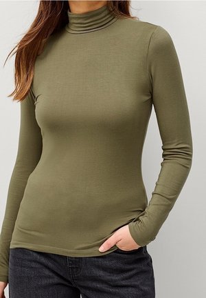 Femme portant un col roulé ajusté à manches longues vert olive, main dans la poche, portant un jean gris foncé, debout devant un fond uni clair.