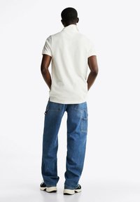 Camisa polo blanca de manga corta, corte relajado, combinada con pantalones cargo de mezclilla azules con bolsillos laterales y una silueta holgada. Zapatillas negras.