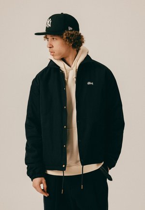 Młody mężczyzna z kręconymi włosami, w czarnej czapce, czarnej kurtce na beżowej bluzie z kapturem, patrzący w lewo na jednolitym tle.