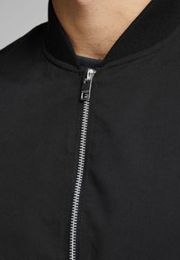 Černá bomber bunda s žebrovaným límcem, se stříbrným zipem a hladkým, texturovaným materiálem. Detailní pohled na výstřih a zip.