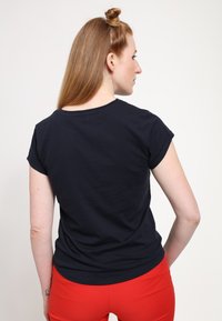 Tyrkysové tričko s krátkým rukávem a kulatým výstřihem, hladký bavlněný materiál, nošeno s červenými skinny kalhotami, pohled zezadu.