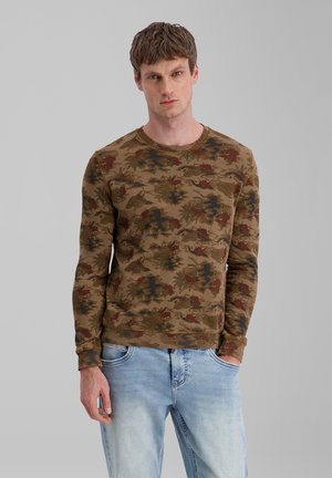 Young man with light skin and short brown hair wearing a brown floral patterned long-sleeve shirt and light blue jeans against a plain background.