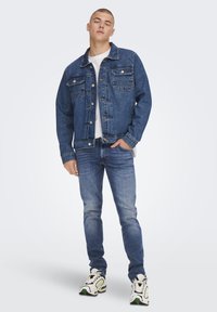 Giubbotto di jeans blu scuro con tasche frontali, chiusura a bottoni, indossato sopra una t-shirt bianca. Jeans azzurri e sneaker bianche con dettagli verdi.