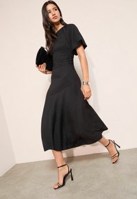 Fekete midi ruha texturált anyagból, rövid pufó ujjakkal és A-vonalú sziluettel; fekete pántos magassarkúval és hozzáillő borítéktáskával párosítva.