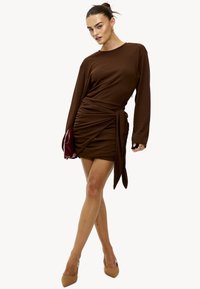 Robe marron à manches longues avec fronces et un détail noué à la taille. Tissu lisse. Associée à un sac à main rouge et des escarpins pointus couleur nude.