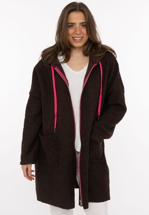 Frau trägt einen langen dunkelbraunen, strukturierten Mantel mit pinkem Reißverschluss und Kordelzug, weißem Hemd und weißer Hose, lächelnd und stehend.