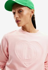 Sudadera rosa con un logo en relieve, acompañada de una gorra verde brillante. Textura suave, con un escote redondo y un corte relajado.