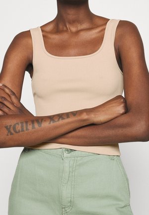 Persona con los brazos cruzados, vistiendo una blusa sin mangas beige y pantalones verde claro, mostrando un tatuaje de números romanos en el antebrazo izquierdo.