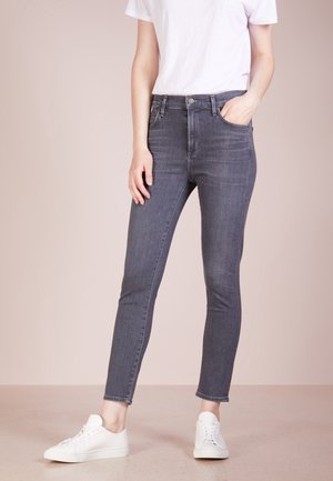 Person iført grå slim fit jeans, hvid t-shirt og hvide sneakers, stående med den ene hånd i lommen mod en ensfarvet baggrund.