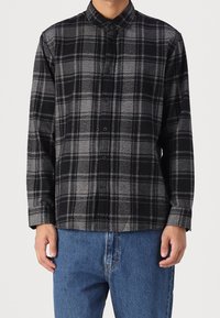 Muž v černé a šedé kostkované košili s límcem a dlouhými rukávy, spárované s modrými džínami.