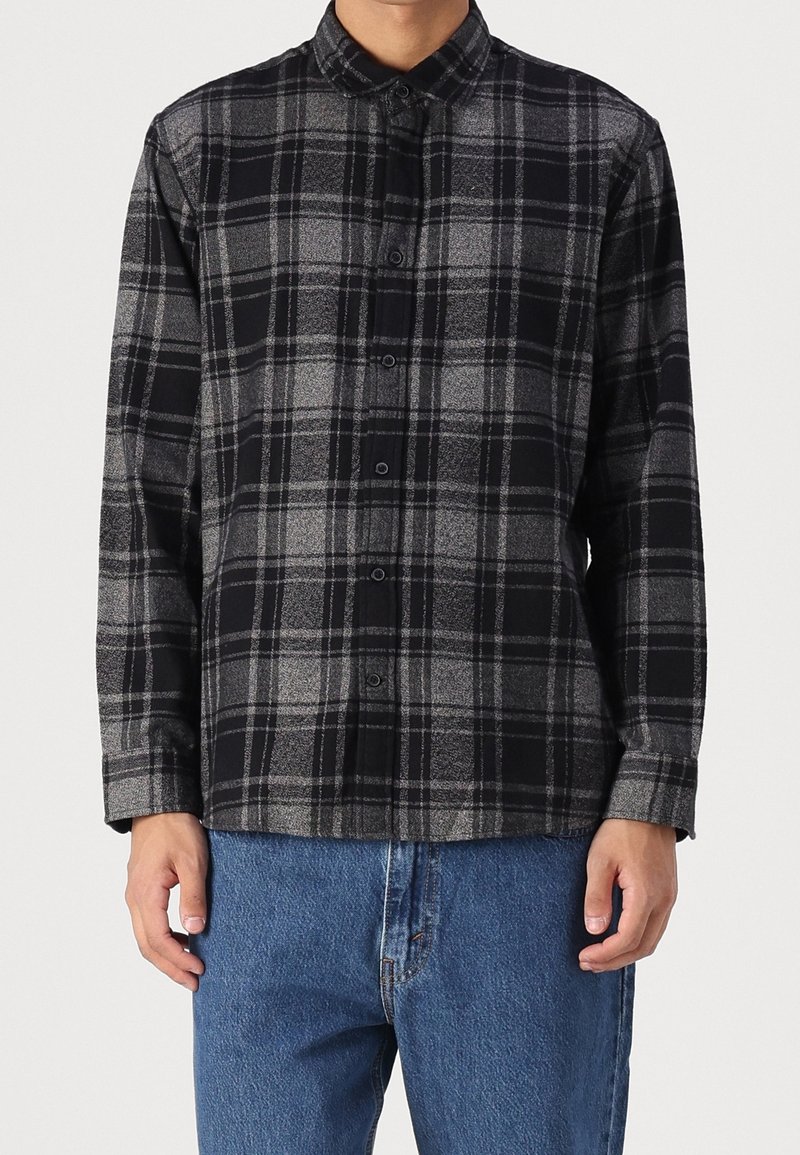 Muž v černé a šedé kostkované košili s límcem a dlouhými rukávy, spárované s modrými džínami.