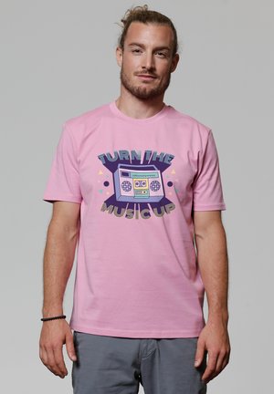 Mann mit leichtem Bart und zurückgebundenen Haaren, der ein pinkes T-Shirt mit einer bunten Kassette und dem Text "DREH DIE MUSIK LAUTER" trägt.