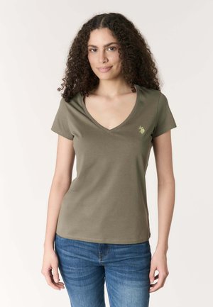 Donna con capelli ricci che indossa una maglietta verde oliva con scollo a V, con un piccolo logo giallo e jeans blu, in piedi davanti a uno sfondo semplice.