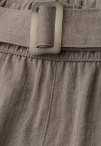 Primer plano de pantalones de tela beige con cinturilla fruncida y cinturón de tela a juego pasado por una hebilla rectangular translúcida.