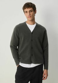 Mörkgrå cardigan i mjukt stickat material. Har V-ringning, knappstängning fram och ribbad mudd. Bärs över en vit skjorta.