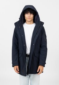 Marineblaue isolierte Parka mit Kapuze, ausgestattet mit einem Reißverschluss und Druckknöpfen, mehreren Taschen und einem strukturierten Stoff mit glattem Futter.