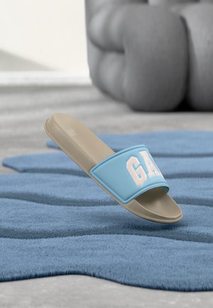 Sandale beige à semelle plate avec une bride bleu clair ornée de grandes lettres blanches "GAP", posée sur un tapis bleu texturé avec un coussin gris flou en arrière-plan.