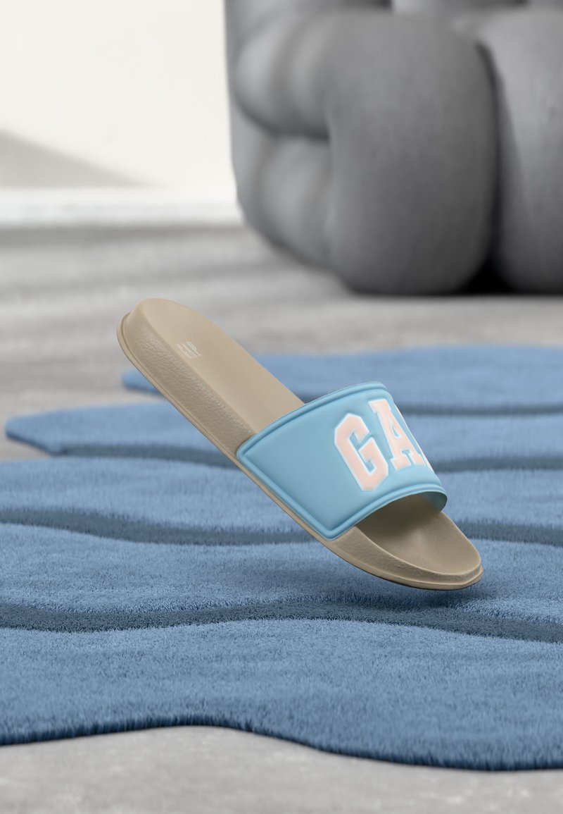 Sandale beige à semelle plate avec une bride bleu clair ornée de grandes lettres blanches "GAP", posée sur un tapis bleu texturé avec un coussin gris flou en arrière-plan.