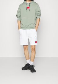 Felpa verde menta con un patch rosso, abbinata a pantaloni corti bianchi con un'etichetta rossa e scarpe da ginnastica nere. Calzini bianchi visibili.