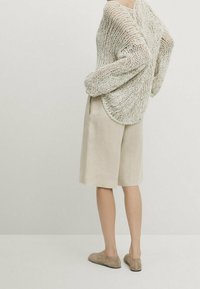 Overmaat gebreide trui in lichtbeige, gecombineerd met beige linnen korte broek en beige instapschoenen, gezien vanaf de achterkant.