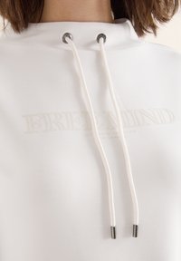 Witte hoodie met een hoge nek, voorzien van een trekkoord met zilverkleurige hardware. "FREE MIND" gedrukt in een subtiele tint op de borst.