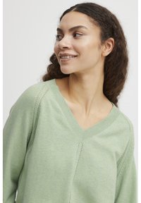 Helles grünes V-Ausschnitt-Pullover mit strukturiertem Strick, der subtile Nahtdetails und eine lockere Passform aufweist. Das weiche, glatte Material sorgt für Komfort.