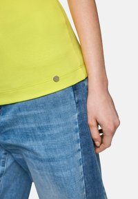 Helles gelbes Baumwoll-T-Shirt mit gerippter Textur und einem silbernen, metallischen Logo-Akzent am Saum, kombiniert mit hellblauen Jeans.