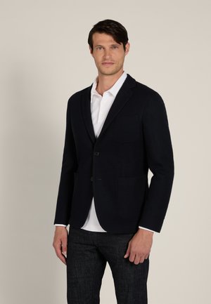 Mann trägt schwarzen Blazer über weißem Poloshirt und dunklen Jeans, steht vor hellem, einfachem Hintergrund.