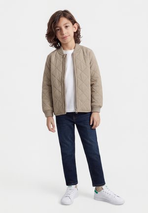 Gyerek áll, bézs színű steppelt dzsekit, fehér pólót, sötétkék farmert és fehér, zöld részletekkel díszített sportcipőt visel, fehér háttéren.