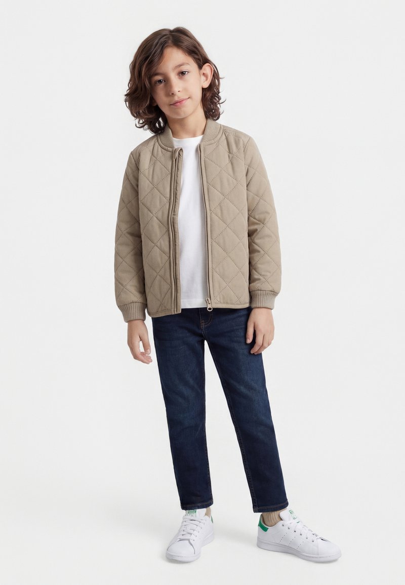 Otrok stoji oblečen v bež prešito jakno, belo majico, temno modre kavbojke in bele superge z zelenimi poudarki na belem ozadju.